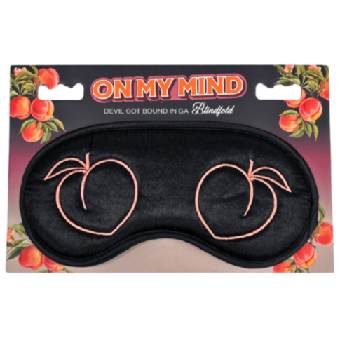 The On My Mind - Devil Got Bound in GA - Blindfold is an elegant black sleep mask with pink embroidered peaches, packaged beautifully in a whimsical peach pattern. The packaging creatively reads On My Mind, echoing the phrase Devil Got Bound in GA.