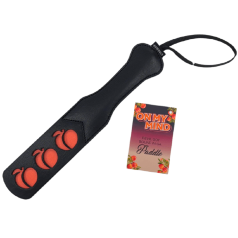A black paddle with a wrist strap features three red peaches. Next to it, a card reads On My Mind - Paddle, highlighted by a sunset gradient with a peach border. This combo echoes the playful mischief of Devil Got Bound from the On My Mind collection.