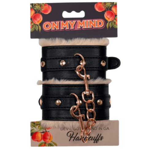 Black leather and fur handcuffs with gold studs and a chain, packaged as On My Mind - Devil Got Bound in GA - Handcuffs featuring peach designs on the label.